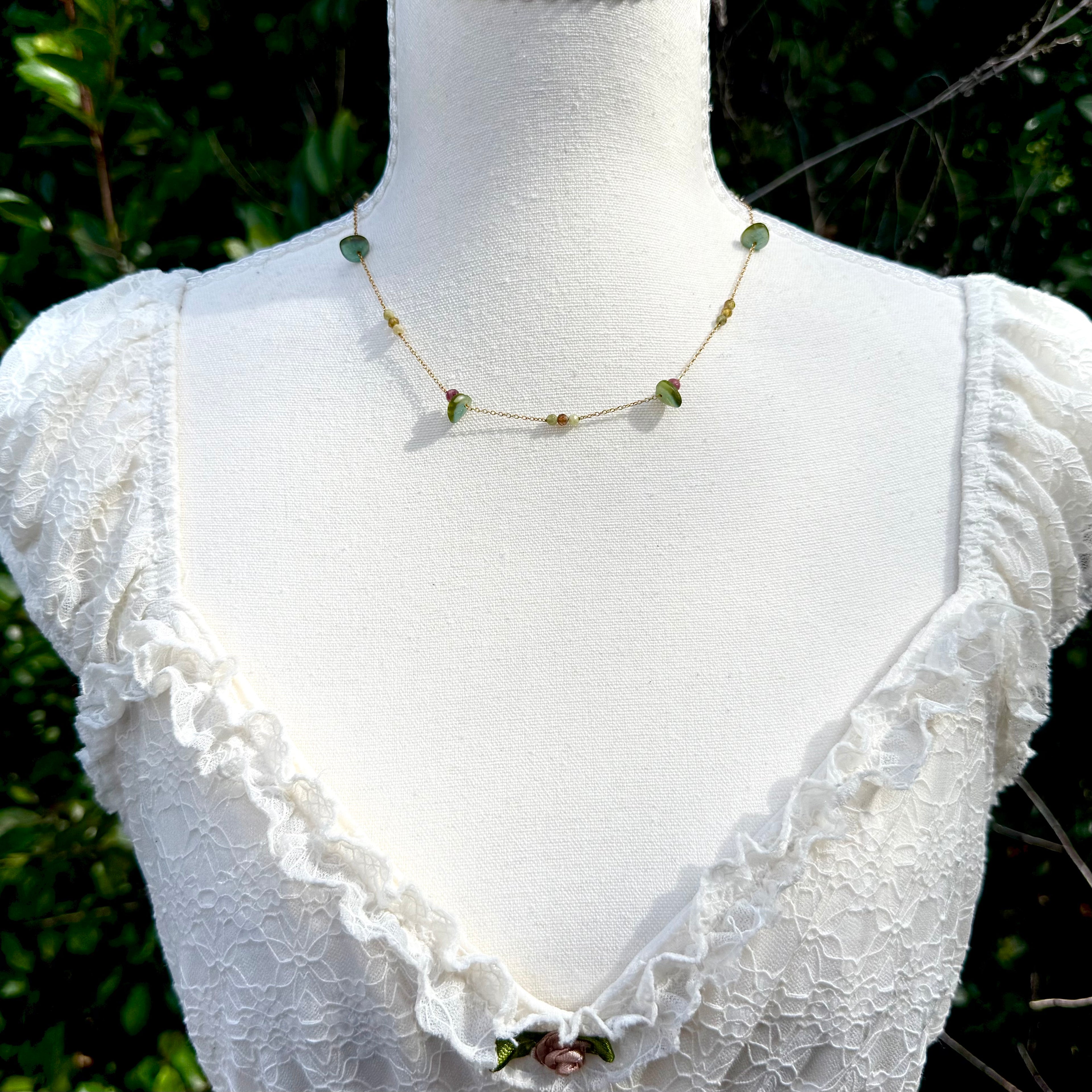 Mannequin wearing a white lace top with a green necklace against a blurred natural background